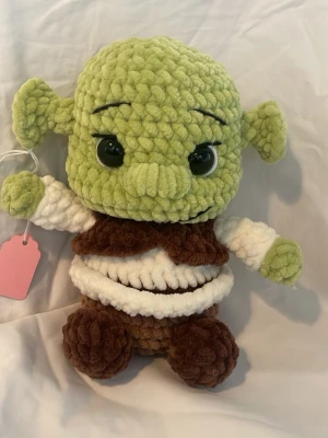Handgjord Shrek-inspirerad mjukisdocka - Supermjuk och gosig Shrek-inspirerad docka, virkad i grönt, brunt och vitt garn. Stora svarta ögon och tydliga ansiktsdrag. Perfekt som present eller dekoration för alla som gillar nördiga prylar och unika gosedjur. 17cm