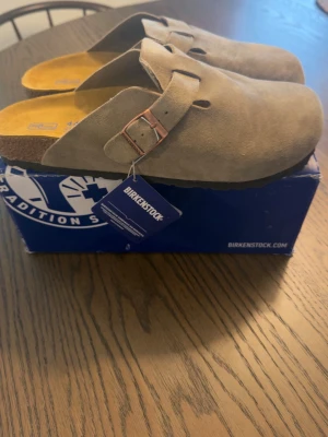 Birkenstock Boston - Säljer ett par snygga beigea Birkenstock tofflor i mjuk mocka med justerbart spänne i metall. Klassisk korksula och ergonomisk fotbädd för extra komfort. Perfekta för chill dagar hemma eller när du vill ha en clean och avslappnad stil.