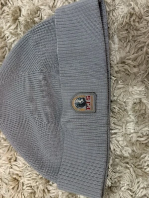 Ljusgrå ribbstickad mössa med patch - Snygg ljusgrå ribbstickad mössa med bred uppvikt kant och en patch framtill med texten 'FJLS' och ett motiv. Perfekt för kalla dagar och enkel att matcha med olika outfits. Materialet känns mjukt och stretchigt.