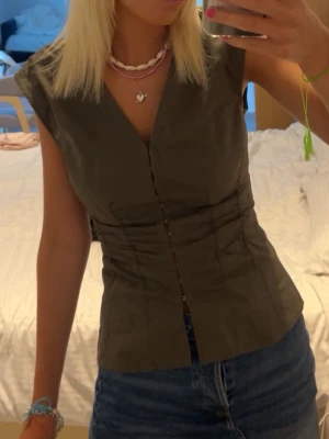 Mörkgrön v-ringad blus - Snygg mörkgrön blus med v-ringning och knappar framtill. Modellen har korta ärmar och figurnära passform som markerar midjan. Perfekt att styla med jeans eller kjol för en trendig look.