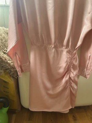 Ljusrosa miniklänning i satin - Supersnygg ljusrosa kortklänning från Les Amourettes Atelier & Other Stories storlek 34 i glansigt satinmaterial. Klänningen har långa ärmar med knapp vid ärmslut, omlottdetalj baktill samt dragkedja och snygg rynkning på sidan med slits fram. Perfekt för dig som vill ha en chic och trendig look eller till bröllop tex.