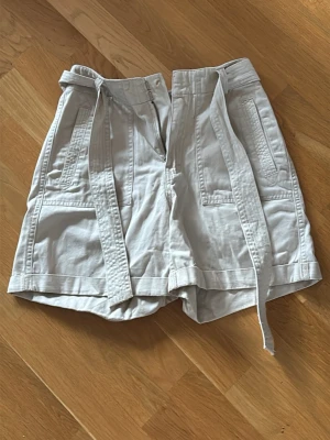 Krämigt Vita högmidjade shorts med knytband - Säljer ett par vita högmidjade shorts med breda ben och knytband i midjan. Shortsen har stora fickor fram och bak, samt dragkedja och knappstängning. Perfekta för varma dagar och en avslappnad stil.