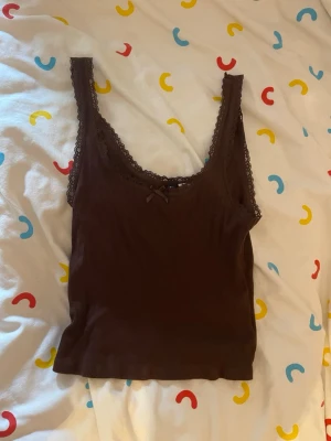Brun ribbad linnetopp från H&M - Söt brun ribbad linnetopp från H&M Divided med spetskant runt urringningen och en liten rosett framtill. Perfekt att styla med jeans eller kjol för en chill look. Materialet är mjukt och stretchigt för extra komfort.