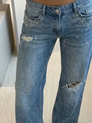 Blå jeans med slitningar och pärldetaljer - Säljer ett par blå jeans med raka ben, slitningar på låret och knät samt vita pärldetaljer över hela framsidan. Jeansen har klassisk femficksmodell och normal midja. Perfekta för dig som vill ha en unik och trendig look. Nypris: 800 SEK
