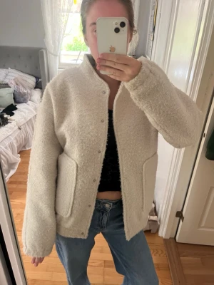 Krämvit teddyjacka med fickor - Mysig krämvit teddyjacka med oversized passform och stora fickor framtill. Jackan har tryckknappar och en hög krage som ger extra värme. Perfekt för kyliga dagar och enkel att styla till jeans eller kjol.