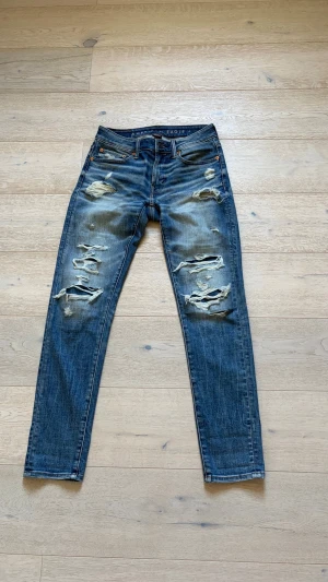 American Eagle jeans  - Säljer ett par blå  jeans med  slitningar och hål på framsidan. Jeansen har klassisk femficksdesign och fadear sjukt snyggt. Perfekta för en avslappnad och trendig streetstil.