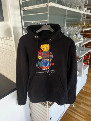 Ralph Lauren Hoodie - Svart hoodie från Ralph Lauren med Polo Bear-tryck framtill. Klassisk känguruficka och justerbar huva med snören. Mjuk bomullskänsla och ribbade muddar vid ärmar och nederkant. Perfekt för en avslappnad och trendig look. Storlek S. Helt ny med prislappen kvar.