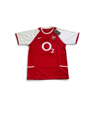 Arsenal Nike matchtröja Henry 14 - Säljer en klassisk Arsenal matchtröja i rött och vitt med tryck 'Henry 14' på ryggen. Tröjan har O2-logga på bröstet, Arsenal-emblem och är tillverkad i lätt och ventilerande material. Perfekt för fotbollsfans och samlare. Kom gärna med bud