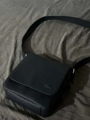 Bag for Salman  - Snygg svart axelväska från Lacoste med klassisk krokodil-logga på framsidan. Väskan har en fyrkantig form, justerbar axelrem och är gjord i slitstarkt material med en stilren look. Perfekt för att bära det viktigaste och matchar enkelt olika outfits.