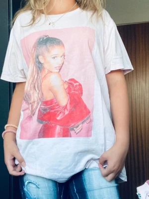 Ariana Grande t-shirt i rosa - Säljer en oversized rosa t-shirt med stort tryck av Ariana Grande på framsidan och turnétext på ryggen. T-shirten har korta ärmar och är gjord i mjuk bomull, perfekt för dig som gillar artistmerch och vill sticka ut. Har några hål, men man ser inte de om man kollar noga 