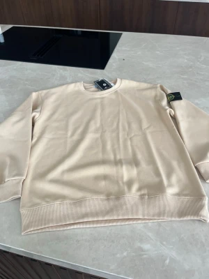 Beige hoodie från Stone Island - Snygg beige hoodie från Stone Island med klassisk logotyp-patch på ärmen. Hoodien har rund halsringning, ribbade muddar och är tillverkad i mjukt bomullsmaterial. Perfekt för en clean och stilren look. Passar även S.