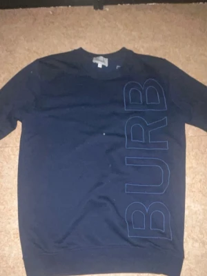 Mörkblå sweatshirt från Burberry - Snygg mörkblå sweatshirt från Burberry med stort tryck 'BURB' i vertikal stil på framsidan. Tröjan har rund halsringning och är tillverkad i mjukt bomullsmaterial. Perfekt för en clean och stilren look.