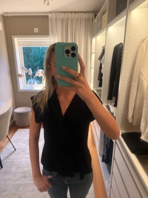 Svart omlottopp med volang - Snygg svart omlottopp med korta ärmar och volangdetalj nedtill. Toppen har en feminin passform och knyts i midjan för en markerad siluett. Perfekt att styla med jeans eller kjol för en trendig look.