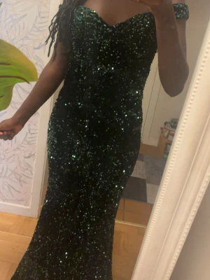 Grön glittrig långklänning FAERIESTY - Supersnygg långklänning från FAERIESTY i mörkgrönt med massor av paljetter. Klänningen har offshoulder-modell och markerad midja som ger en elegant siluett. Perfekt för dig som vill sticka ut med en glittrig look.