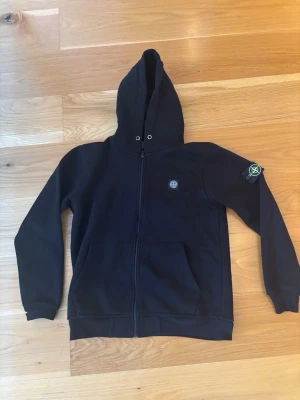 Svart hoodie från Stone Island - Säljer en svart hoodie från Stone Island med dragkedja framtill och klassisk logotyp på vänster ärm. Hoodien har huva med dragsko och två fickor framtill. Perfekt för dig som gillar streetwear och vill ha en stilren look.