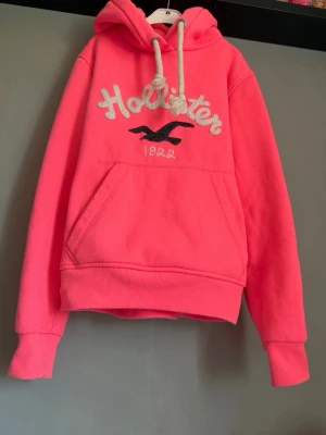 Rosa hoodie från Hollister med huva - Snygg rosa hoodie från Hollister med stor magficka, broderad logga och text på bröstet samt vita dragsnören i huvan. Tillverkad i mjukt material som känns skönt mot huden. Perfekt för en avslappnad och trendig look.