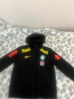 Svart Brasil Nike träningsjacka - Säljer en svart träningsjacka med huva från Nike, dekorerad med Brasilien fotbollslandslagets logga och sponsorer i gult och rött. Jackan har dragkedja framtill och tryck på både bröst och ärmar. Perfekt för dig som gillar fotboll och vill ha en sportig look.
