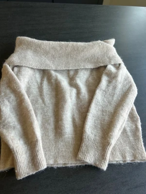 Beige off shoulder stickad tröja - Mysig beige stickad tröja med bred ribbad off shoulder-krage och långa ärmar. Tröjan har ribbade muddar vid ärmslut och nederkant, vilket ger en snygg och avslappnad look. Perfekt för dig som gillar en enkel men trendig stil.