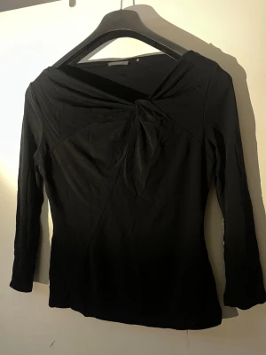 Svart långärmad topp från Tahari - Snygg svart långärmad topp från Tahari med twistad detalj vid halsringningen. Toppen har en smickrande passform och är tillverkad i ett mjukt material som känns bekvämt mot huden. Perfekt att styla med jeans eller kjol för en trendig look.