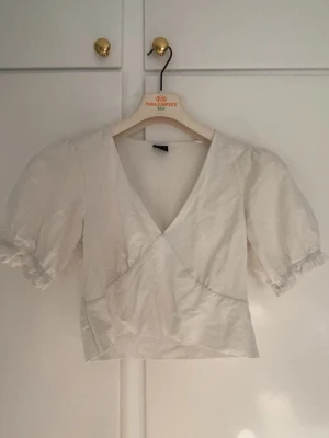 Vit omlottopp från Gina Tricot - Säljer en söt vit omlottopp från Gina Tricot med puffärmar och v-ringning. Toppen har korta ärmar med resår och volangdetalj vid ärmslut. Perfekt för dig som gillar en feminin och trendig stil.
