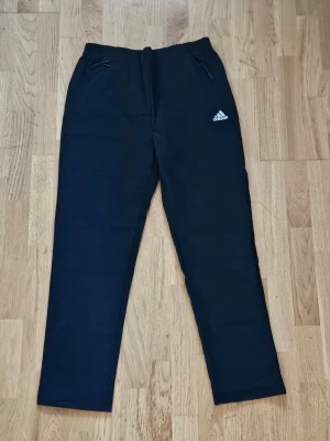 Svarta mjukisbyxor från Adidas - Svarta mjukisbyxor från Adidas med raka ben och dragsko i midjan. Byxorna har två sidofickor med dragkedja och Adidas-logga på vänster lår. Perfekta för chill eller träning. Tillverkade i mjukt material för maximal komfort.