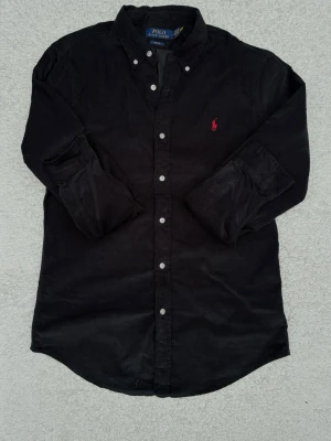 Polo Ralph Lauren Manchester Skjorta - Polo Ralph Lauren Manchester Skjorta – Slim Fit – Strl M  Säljer en svart Polo Ralph Lauren manchesterskjorta i storlek M (Slim Fit). Klassisk modell med röd broderad logga på bröstet och button down-krage.  Skjortan är i fint använt skick utan hål eller fläckar. Perfekt till både vardag och mer uppklädda tillfällen.  - Storlek: M (Slim Fit) - Färg: Svart - Äkta Polo Ralph Lauren Hör gärna av dig om du vill ha mått eller fler bilder.