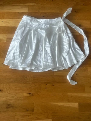 Vit omlottkjol i satin - Säljer en vit omlottkjol i glansigt satinmaterial med knytband i midjan. Kjolen har en kort och luftig passform som ger en snygg siluett.