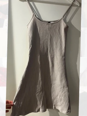Beige ribbad kort klänning - Säljer en beige ribbad kort klänning med smala axelband och rundad halsringning. Klänningen har en figurnära passform och är tillverkad i ett mjukt, stretchigt material som passar perfekt till sommaren.