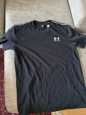 Svart Under Armour t-shirt - Snygg svart t-shirt från Under Armour med logga på bröstet och textdetaljer längs axlarna. Klassisk rund hals och korta ärmar. Perfekt för dig som gillar sportig stil och enkel design.