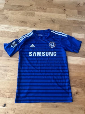 Chelsea FC blå matchtröja Adidas - Chelsea FC matchtröja i blått med vita detaljer och horisontella ränder. Adidas-logga på bröstet, klubbmärke och Samsung som sponsor framtill. Korta ärmar med Premier League-märke på ena ärmen och klassiska Adidas-ränder på axlarna.