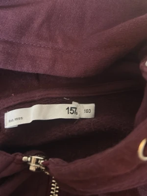 Vinröd hoodie med dragkedja - Mysig vinröd hoodie med dragkedja framtill och två stora fickor. Tröjan har huva med dragsko och ribbade muddar vid ärmslut och nederkant. Perfekt för chill dagar eller när du vill ha något bekvämt och snyggt. rätt så stor i storleken