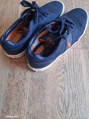 Mörkblå Polo Ralph Lauren sneakers - Snygga mörkblå sneakers från Polo Ralph Lauren med klassisk låg profil. Skorna har vita sulor med orange detalj och den ikoniska loggan på sidan. Ovandelen är i slätt material och insidan har en kontrasterande brun ton. Perfekta för en stilren och avslappnad look.