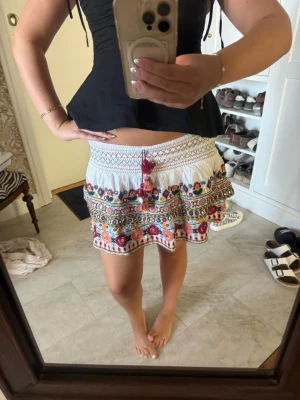 Vit broderad kortkjol från Zara - Supersöt vit kortkjol från Zara med färgglada broderier och volangkant. Kjolen har snörning med tofsar i midjan och detaljer i rött, blått, gult och grönt. Perfekt för sommarens alla äventyr och ger en bohemisk vibe.
