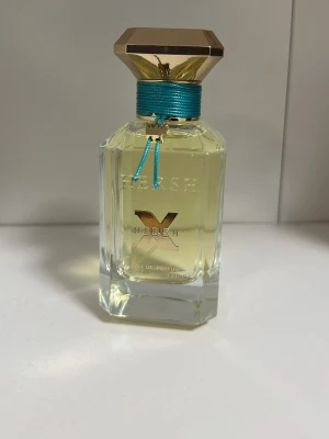 Hersh x av hers parfym - Snygg parfymflaska i genomskinligt glas med guldfärgat lock och turkost band runt halsen. Flaskan har en elegant, kantig form och detaljer i guld. Texten 'X HER&SH' syns på framsidan. Perfekt för dig som gillar exklusiv design.