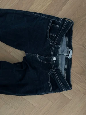 Mörkblå bootcut jeans med kontrastsömmar - Säljer ett par mörkblå bootcut jeans med vita kontrastsömmar och klassisk. Jeansen har låg midja och utsvängda ben. Knappt använda