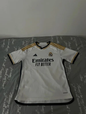 Real Madrid matchtröja Adidas vit/guld - Säljer en Real Madrid matchtröja från Adidas i vitt med guldiga axelränder och svarta detaljer. Tröjan har klubbmärke och sponsortryck på bröstet samt korta ärmar. Perfekt för dig som älskar fotboll och vill representera ditt favoritlag.