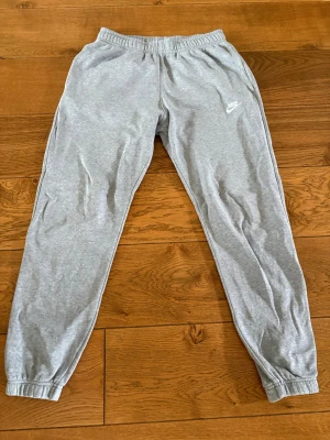 Gråa mjukisbyxor Nike - Snygga gråa mjukisbyxor från Nike med klassisk logga på vänster lår. Byxorna har resår i midjan och vid bensluten för en skön passform. Tillverkade i mjukt bomullsmaterial som är perfekt för chill eller träning. 