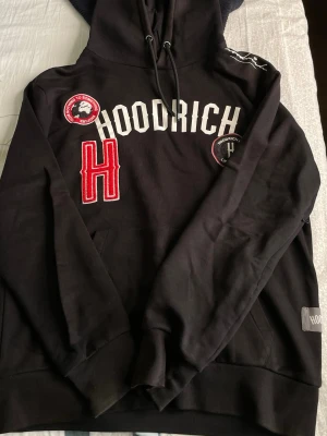 Svart hoodie från Hoodrich - Svart hoodie med broderade patches och tryck från Hoodrich. Stor vit logga framtill, röd H-detalj och flera märken på bröstet. Klassisk huva med dragsko och känguruficka. Perfekt för en streetwear-look.