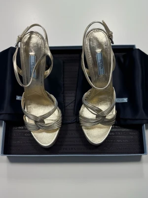 Prada Sandaler Guld Metallic Heels / EU 39,5 / Oanvända i originalbox - Medföljer: originalbox från Prada samt dustbags. Oanvända Prada-sandaler i guldig metallic nappa-läder med kvarvarande prislapp. Sulorna är helt orörda. Samtliga originalhandlingar och förpackningar medföljer.  Elegant design med flätade/knutna metallic-remmar vid tån, smal ankelrem med silvertonerat spänne och hög stilettklack. Pradas triangellogotyp applicerad på sulan.  Märke: Prada Milano 