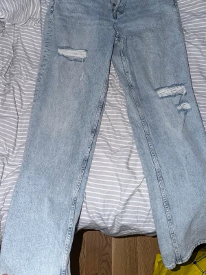 Ljusblå raka jeans med slitningar - Säljer ett par ljusblå jeans med raka ben och trendiga slitningar på båda benen. Jeansen har hög midja och knäppning framtill. Perfekta för en avslappnad och cool stil. 