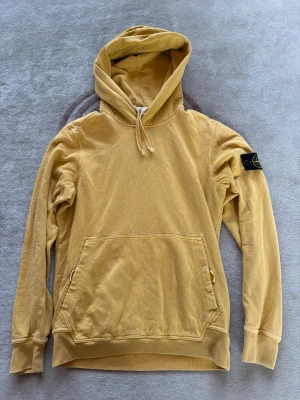 Gul hoodie från Stone Island - Säljer en gul hoodie från Stone Island med klassisk patch på vänster ärm. Hoodien har dragsko i huvan och en stor magficka framtill. Tillverkad i mjuk bomull och har ribbade muddar vid ärmar och nederkant. Perfekt för en avslappnad och trendig stil.