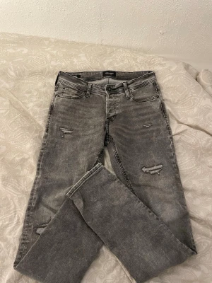 Grå slitna jeans från Jack & Jones - Säljer ett par grå slim fit jeans från Jack & Jones med slitningar och trasiga detaljer på benen. Jeansen har klassisk femficksmodell och knappgylf. Perfekta för en avslappnad och trendig look.