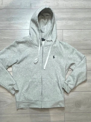  Ralph Lauren Hoodie / Tröja - Ljusgrå hoodie med dragkedja och huva, justerbara snören och fickor framtill. Klassisk passform och mjukt material, perfekt för en avslappnad stil. Liten broderad logga på bröstet ger en clean look.