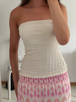 Vit bandeau tubtopp - Säljer en stilren vit bandeau tubtopp i smockat tyg. Toppen är ärmlös och har en tight passform som sitter snyggt på kroppen. Perfekt att matcha med kjol eller jeans för en fräsch look.