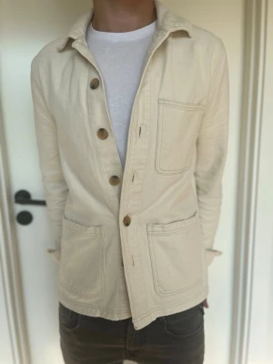 Ljusbeige overshirt med stora knappar - Säljer en stilren ljusbeige overshirt i kraftigt bomullstyg. Jackan har tre stora utanpåfickor, klassisk krage och stängs med fem bruna knappar. Perfekt lager-på-lager-plagg för dig som gillar enkel och clean stil. Overshirten har några små fläckar men inget man tänker på. Modellen är 179cm lång