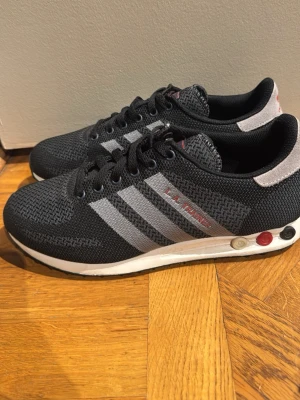 Adidas la trainer skor  - Snygga svarta sneakers från adidas med grå ränder på sidorna och vita detaljer på sulan. Skorna har en stickad ovandel i textil och klassisk snörning. Sulan har färgade detaljer och ger en sportig look. Perfekta för dig som gillar streetwear och enkel stil.