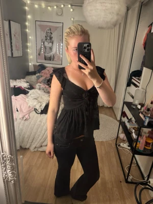 Svart blus med volang och v-ringning - Säljer en svart blus med v-ringad hals och volangdetaljer vid axlarna. Toppen har en lätt rynkad effekt framtill och är något utsvängd nedtill. Perfekt för dig som gillar en feminin och trendig stil.