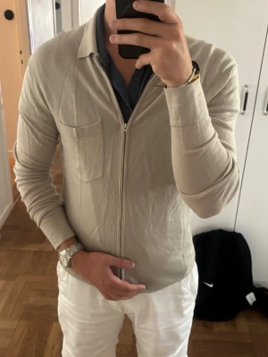 Beige ziptröja Merinoull JL - Snygg beige ziptröja med lång ärm och bröstficka. Tröjan har en smal passform och är tillverkad i ett tunt, mjukt material som passar perfekt till sommaren. Enkel och stilren design utan synligt märke. Perfekt att slänga på över en t-shirt eller skjorta. Jag har medium i allt och så sitter den på mig.