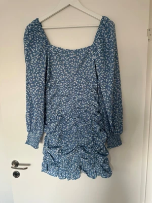 Blå blommig kortklänning med volang - Supersöt blå kortklänning med vitt blommönster och volangdetaljer. Klänningen har långärmade puffärmar med smock vid ärmslut och en fyrkantig ringning. Perfekt för sommarens alla tillfällen!