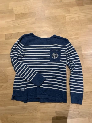 Blåvit randig långärmad Morris tröja - Snygg blå Morris tröja med vita horisontella ränder och en bröstficka med broderad logga. Tröjan har rund halsringning och långa ärmar. Perfekt för en avslappnad och stilren look.